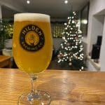 A half pint of Hilden Brewery's Belfast Blonds sits on the bar top with a Christmas Tree in the background all lit up.