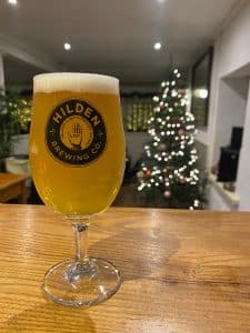 A half pint of Hilden Brewery's Belfast Blonds sits on the bar top with a Christmas Tree in the background all lit up.